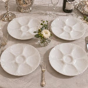 Puede incluir: Cuatro platos de ostras blancos con seis compartimentos cada uno, dispuestos sobre una mesa preparada para una cena formal. También hay cubiertos de plata, copas de vino y un pequeño arreglo floral. Los platos tienen un borde festoneado.