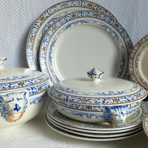 Peut inclure: Ensemble de vaisselle en porcelaine blanche avec des motifs décoratifs bleus et dorés. L'ensemble comprend des assiettes, un plat de service couvert et une soupière avec des poignées en tête de lion. Le design présente des motifs floraux et de volutes.