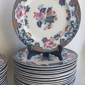 Può includere: Una grande pila di piatti rotondi in ceramica con un motivo floreale centrale nei toni del rosa, arancione e blu scuro. L'ampio bordo è decorato con un motivo blu scuro e oro. Un piatto è esposto in verticale su un supporto in plastica nera.
