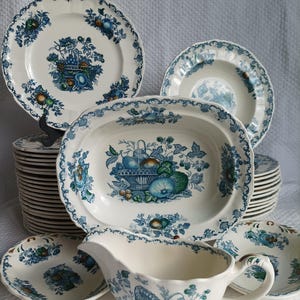 Peut inclure: Ensemble de vaisselle en céramique blanche avec un motif de panier de fruits bleu. L'ensemble comprend des assiettes, des bols, un plat de service et une saucière. Les bords des assiettes et des bols sont festonnés.