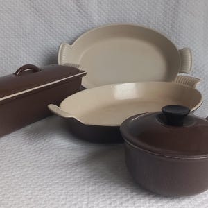Puede incluir: Colección de utensilios de cocina de cerámica marrón y crema. Incluye un molde rectangular para pan con tapa, dos fuentes ovaladas y una olla redonda pequeña con tapa y asa. Los utensilios están sobre una superficie blanca texturizada.