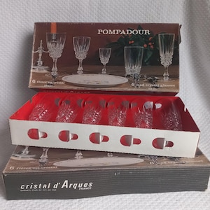 Peut inclure: Deux boîtes de verrerie en cristal vintage. La boîte supérieure est étiquetée "POMPADOUR" et contient divers verres en cristal et une assiette. La boîte inférieure est étiquetée "cristal d'Arques FRANCE". Les verres sont clairs et ornés.