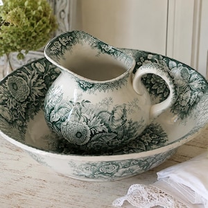 May include: A ceramic wash basin and matching pitcher set featuring a cream-coloured base with a detailed dark green floral transferware pattern. The pitcher has a curved handle and a wide spout, while the basin is large and circular.