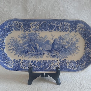 Peut inclure: Un plat de service ovale en céramique avec un motif bleu et blanc. Le centre représente un paysage avec des arbres et un petit bâtiment, tandis que la large bordure présente des motifs floraux complexes et des volutes décoratives.