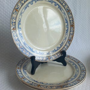 Peut inclure: Trois assiettes en céramique blanc cassé avec une bordure décorative. La bordure présente des motifs floraux et géométriques bleus, dorés et orange. Les assiettes sont présentées sur un support noir sur fond blanc.