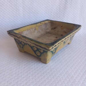 Op de afbeelding: Een rechthoekige keramische bonsaipot met een geel geglazuurde buitenkant voorzien van donkerblauwe decoratieve lijnpatronen en schulpmotieven. De pot heeft een brede rand, vier korte pootjes en een lichtbeige, ongeglazuurde binnenkant.