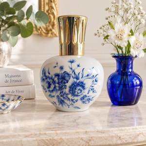 May include: A white ceramic fragrance lamp with a rounded body featuring a blue floral pattern and a gold-toned metal crown-shaped top. The lamp has a smooth, glossy finish and sits as a decorative object with a distinct neck and base design.