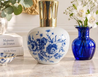 Könnte beinhalten: Eine weiße Duftlampe aus Keramik mit einem abgerundeten Körper, der ein blaues Blumenmuster aufweist, und einem goldfarbenen, kronenförmigen Metallaufsatz. Die Lampe hat eine glatte, glänzende Oberfläche und dient als dekoratives Objekt.