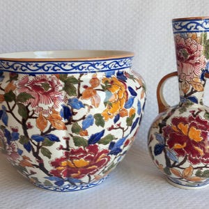 May include: A ceramic planter and a matching two-handled vase, both featuring a cream-colored base with a vibrant, multi-colored floral pattern of red, yellow, and blue peonies, accented by a blue decorative border along the rims and base.