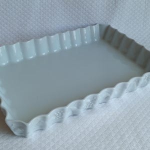 Könnte beinhalten: Eine rechteckige weiße Auflaufform aus Keramik mit glänzender Oberfläche und einem gewellten, geriffelten Rand. Die Außenseiten der Form weisen ein dezentes, geprägtes Muster aus kleinen Traubenclustern auf, das dem Rand Struktur verleiht.