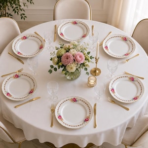 Puede incluir: Una mesa redonda preparada para una cena formal con platos blancos con un borde floral. Cubiertos dorados, copas de cristal y un centro de mesa floral completan el elegante entorno. La mesa está cubierta con un mantel blanco.