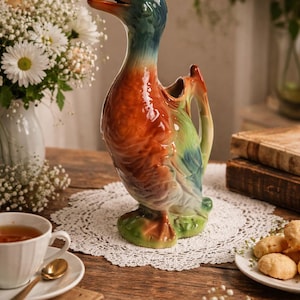 Op de afbeelding: Een keramische eendvormige kan met een levendig kleurenschema van oranje, groen en blauw. De kan heeft een handvat en een tuit en staat op een houten tafel met een witte kanten placemat. Een kopje thee en een bord met koekjes staan ook op de tafel.