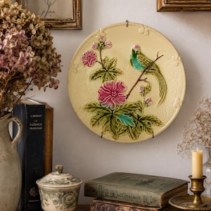 Könnte beinhalten: Ein runder, cremefarbener Keramik-Wandteller mit einem erhabenen, dreidimensionalen Reliefdesign eines Vogels, der auf einem Ast mit rosa Blüten und grünen Blättern sitzt. Der Teller hat einen gewellten Rand und ist mit Metallclips befestigt.