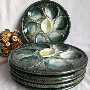 Pode incluir: Uma pilha de seis pratos de ostras de cerâmica verde, cada um com seis poços individuais e um poço central para o molho. Os pratos têm uma superfície texturizada e uma borda em forma de corda. Os pratos são de cor verde escuro com detalhes em verde claro e branco.