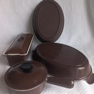 May include: A collection of brown Le Creuset stoneware including a rectangular lidded terrine, a round lidded saucepan with a long handle, and an oval baking dish. The oval dish features the number 28 and the text Made in France on the base.