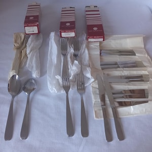 Puede incluir: Tres cajas de cubiertos Guy Degrenne, con el texto "Guy Degrenne" impreso en la parte delantera. Las cajas son rojas y blancas. También hay varias piezas de cubiertos dispuestas sobre una superficie blanca, incluyendo tenedores, cucharas y cuchillos.