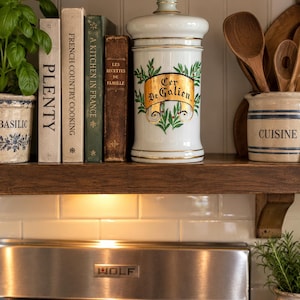 May include: A white ceramic apothecary jar with a lid, featuring a gold-trimmed label with the text Cer De Galien and green leaf illustrations. The jar is positioned on a wooden shelf next to several books and kitchen crocks with text.