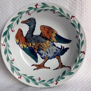 May include: White ceramic plate with a hand-painted duck design in shades of blue, orange, and yellow. The plate features a green and red berry wreath border. A decorative piece for kitchen or dining.