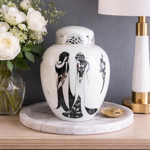 Op de afbeelding: Witte keramische gemberpot met deksel, versierd met zwart-wit illustraties van figuren in traditionele kleding. De pot staat op een marmeren dienblad, met een boeket witte rozen in de buurt.