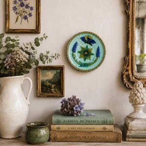 Puede incluir: Un plato decorativo de cerámica con una flor central verde en forma de estrella, tallos florales azules y una mariposa azul sobre un fondo azul claro texturizado, expuesto junto a libros antiguos, una jarra blanca y cuadros.