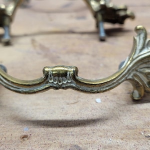 May include: Ornate brass drawer pulls with a decorative leaf design. The pulls have a curved shape with a central decorative element and are mounted on small metal posts. The brass has a slightly aged appearance.
