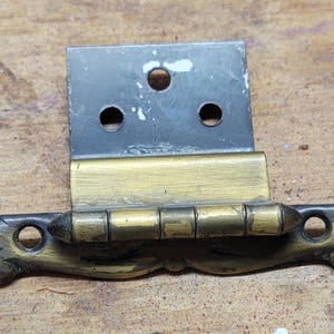 May include: A vintage cabinet hinge with a decorative design. The hinge features a rectangular black plate with three holes, a gold-colored central section, and ornate, curved ends. The hinge is likely made of metal.