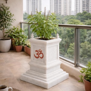 Puede incluir: Una jardinera de mármol blanco con un símbolo Om rojo, que contiene una planta verde con flores moradas. La jardinera está en un balcón con vistas a la ciudad. La jardinera mide aproximadamente 61 cm de altura.