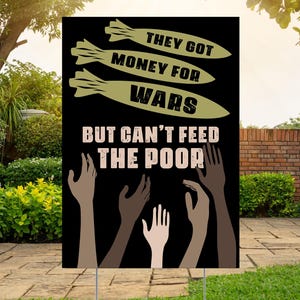 May include: A black yard sign with the text "THEY GOT MONEY FOR WARS BUT CAN'T FEED THE POOR." The sign features illustrations of bombs and hands reaching upwards, set against a backdrop of green grass and a brick wall.