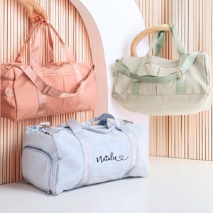 May include: Three duffel bags in pastel colors: peach, mint green, and light blue. The peach bag is held by a hand. The light blue bag has the name "Natalie" printed on it. Each bag has a shoulder strap and a top handle.