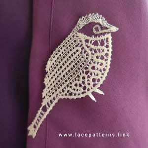 May include: A delicate white lace bird design, intricately crafted with a detailed pattern. The lace is displayed on a purple background.