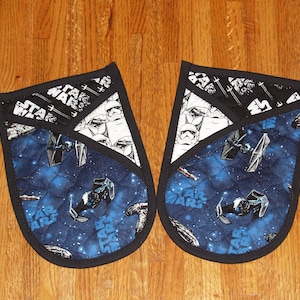 May include: A pair of black oven mitts with a blue and white Star Wars themed fabric design. The fabric features a galaxy pattern with starships and the words "Star Wars".