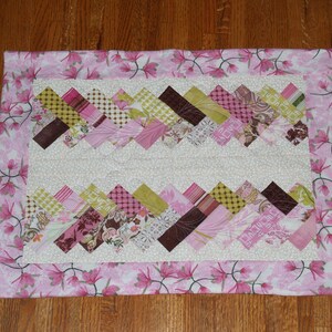 May include: A pink and green patchwork quilt with a white background and a floral border. The quilt is made up of squares of different fabrics, and the border is made of a pink fabric with white flowers.