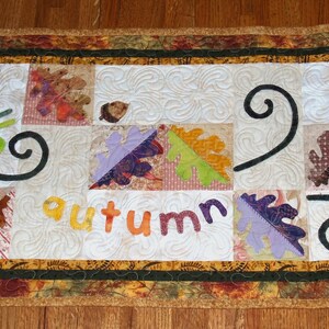 Può includere: Un runner da tavolo bianco trapuntato con un bordo marrone, verde e dorato. Il runner presenta un design a patchwork con varie forme di foglie e le parole "hello autumn".
