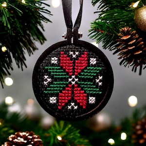 May include: A black circular ornament with a red and green cross-stitch design. The ornament is hanging from a black ribbon and is surrounded by a green Christmas tree.