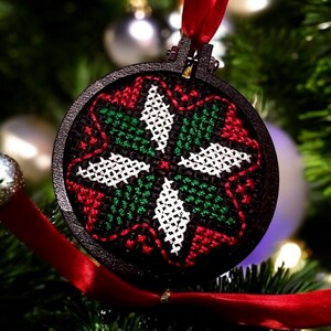 May include: A round, black Christmas ornament with a cross-stitch design in red, green, and white. The ornament is suspended by a red ribbon and hangs on a Christmas tree with silver ornaments.