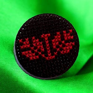 Puede incluir: Un cojín de alfileres circular negro con un diseño bordado en rojo de la palabra "SUCK" en mayúsculas.