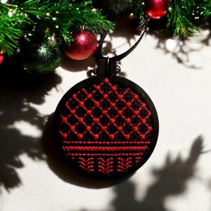 以下が含まれることがあります： 黒と赤の刺繍が施された円形のクリスマスオーナメント。オーナメントは、黒い背景に赤いステッチで幾何学模様が施されています。