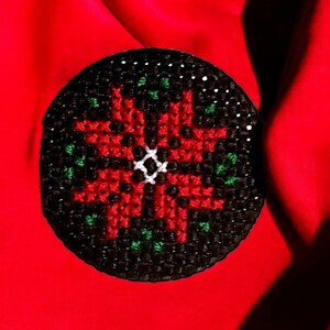 May include: A red and green cross-stitch ornament featuring a poinsettia flower design. The ornament is circular and has a black background.