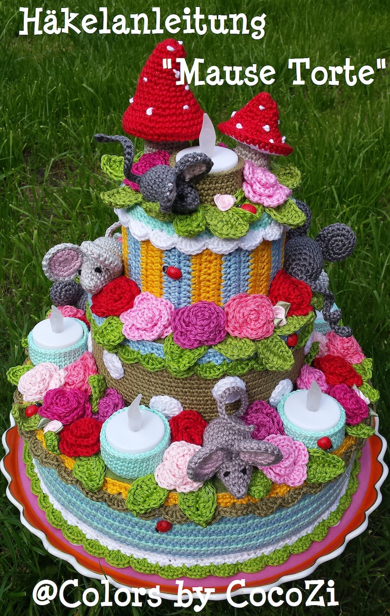 May include: A crocheted cake with three tiers decorated with flowers, leaves, and small mice. The cake is topped with two red mushrooms and a tea light. The cake is on a plate with a scalloped edge. The text "H&auml;ckelanleitung "Maus Torte"" is at the top of the image.