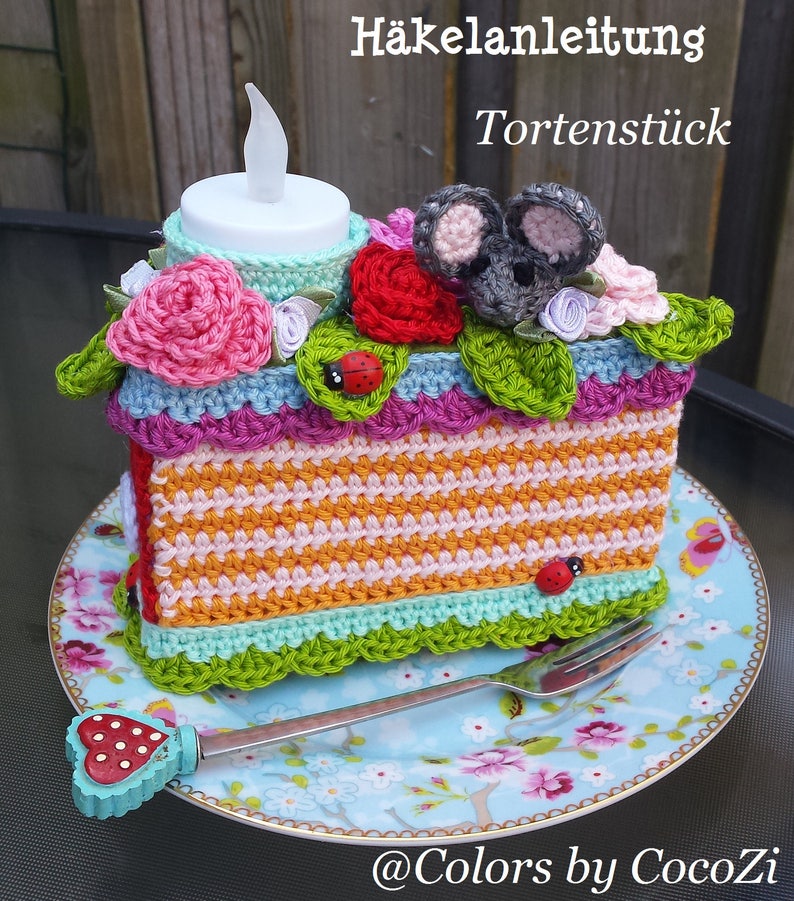 May include: Crocheted cake slice with a gray mouse, red and pink flowers, and a teal heart. The cake slice is on a blue and white floral plate with a fork. The text "H&auml;kelanleitung Tortenst&uuml;ck" is above the cake slice.