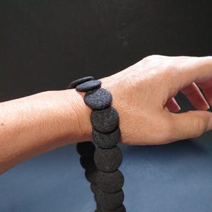 Könnte beinhalten: Ein schwarzes Armband aus Lavasteinen mit runden Perlen. Das Armband wird am Handgelenk getragen.