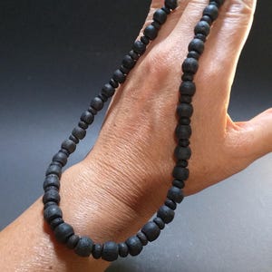 Peut inclure: Un collier de perles noires composé de perles rondes, légèrement irrégulières. Le collier est présenté sur le poignet d'une personne.
