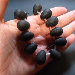 May include: A strand of black lava rock beads, strung on clear thread. The beads are oval shaped and have a rough, textured surface.