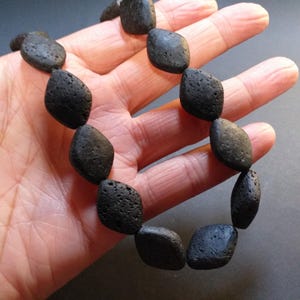 May include: A black stone necklace featuring irregularly shaped, dark gray beads. The beads have a rough, textured surface and are strung together to form a necklace. The necklace is held in a hand.