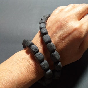 Op de afbeelding: Twee zwarte kralenarmbanden gemaakt van lavasteen. De armbanden zijn gestapeld op de pols van een persoon.