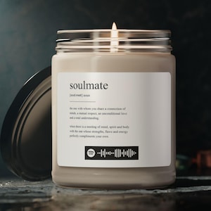 May include: A lit candle in a glass jar with a silver lid. The label reads "soulmate" with a definition and a Spotify code. The candle is on a dark, reflective surface.