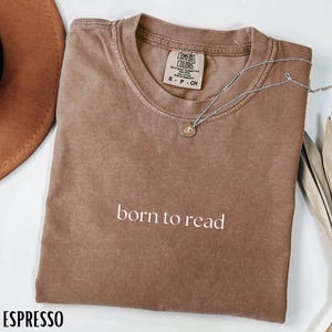 May include: A brown short-sleeved t-shirt with the text born to read printed in white on the chest. The shirt features a Comfort Colors brand label at the inner neckline and is paired with a delicate silver chain necklace with a small round pendant.