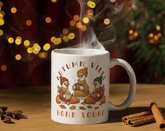 May include: White ceramic mug with the words "AUTUMN VIBES" and "HONK SQUAD" printed on it. The mug features a cartoon illustration of three geese wearing hats and scarves, with pumpkins and fall leaves.
