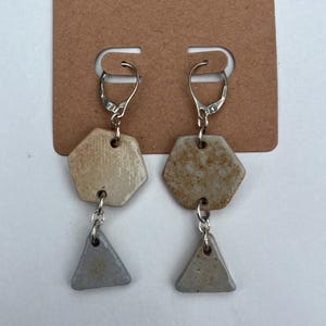 Könnte beinhalten: Ein Paar Ohrhänger mit silberfarbenen Klappbügeln. Jeder Ohrring besteht aus einem sechseckigen Keramik-Anhänger, der durch einen silbernen Ring mit einem kleineren dreieckigen Keramik-Anhänger verbunden ist. Gesprenkelte, erdige Oberfläche.