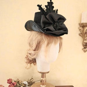 May include: A black hat with a wide brim and a large black bow on top. The hat is decorated with black feathers and a small black jewel.